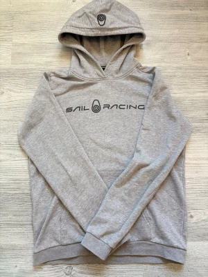 Grå hoodie från Sail Racing - Helt ny grå hoodie från Sail Racing med svart logga tryckt över bröstet och en mindre logga på huvan. Klassisk passform med känguruficka framtill och mjukt material som känns skönt mot huden. Perfekt för en avslappnad och sportig stil.