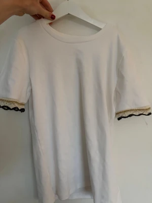 Vit t-shirt med spetsdetalj från Zara - Säljer en vit t-shirt från Zara med korta ärmar och snygga spetsdetaljer i svart och guld vid ärmsluten. T-shirten har en klassisk rund halsringning och är gjord i mjuk bomull. Perfekt för dig som vill ha något enkelt men ändå med en twist. 
