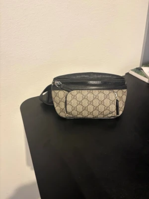 GG  beige magväska - Snygg magväska från GG i beige canvas med klassiskt GG-monogram och svarta detaljer i läder. Väskan har två fack med dragkedja och justerbart band. Perfekt för dig som vill ha en trendig accessoar.