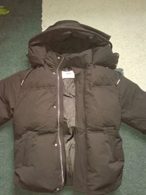 Svart pufferjacka från Ami - Säljer en svart pufferjacka från Ami Alexandre Mattiussi med stor huva och dragkedja framtill. Jackan har två stora fickor och en clean look som passar perfekt till kalla vinterdagar. Materialet är slitstarkt och håller värmen riktigt bra. Köparen får även en säck med sig som kan användas på många sätt, utan att det kostar något extra!