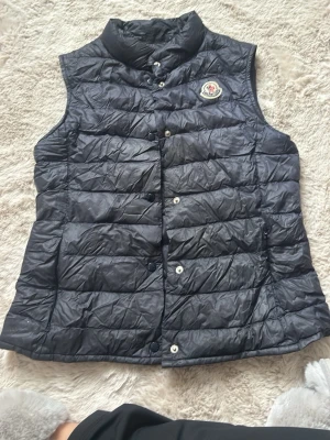 Svart dunväst från Moncler - Snygg svart dunväst från Moncler med klassisk quiltad design och hög krage. Västen har tryckknappar framtill och det ikoniska Moncler-märket på bröstet. Perfekt att slänga över en hoodie eller tröja för extra värme och stil.