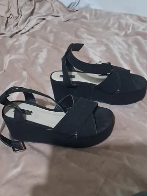 Svarta platåsandaler från NLY SHOES - Snygga svarta sandaler med platåsula från NLY SHOES. Modellen har breda korslagda remmar framtill och justerbar rem runt ankeln med spänne. Perfekta för dig som gillar en stilren look med lite extra höjd. Materialet är textil mocka och sulan är i syntet.