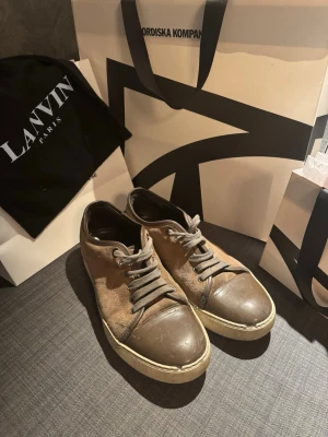 Bruna sneakers från Lanvin - Snygga bruna sneakers från Lanvin med grå snörning. Ovandelen är i mocka. Yttersulan är vit och ger en klassisk kontrast. Perfekta för dig som gillar stilrena och exklusiva sneakers. Värt att tillägga är att lanvin påsen tillhör skorna 