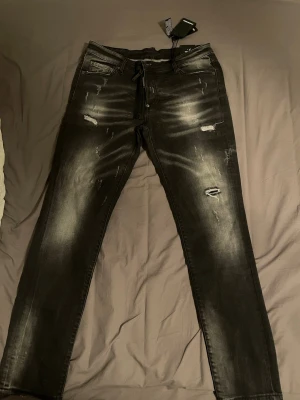 Svarta slitna jeans D2squared - Säljer ett par svarta jeans med slitningar och snygga tvättade detaljer. Jeansen har snörning i midjan istället för klassisk knapp och dragkedja, samt fem fickor. Materialet är stretchigt denim som ger en bekväm passform. Perfekta för en trendig och avslappnad stil.