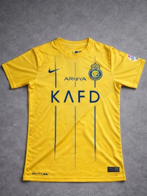 Al Nassr fotbollströja  - Snygg gul matchtröja från Al Nassr med blå logga och tryck. Tröjan har korta ärmar, rund hals och är tillverkad i lätt, ventilerande Dri-FIT-material. Perfekt för fotbollsträning eller att bära som supporter.