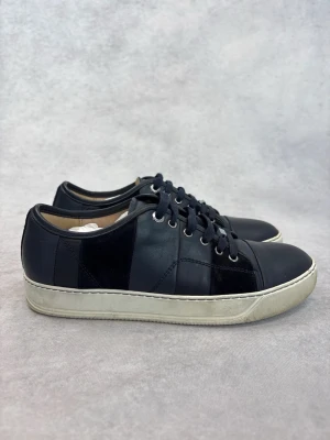 Svarta lädersneakers från Lanvin - Stilrena svarta sneakers från Lanvin i läder med tonade paneler och klassisk vit sula. Skorna har svarta skosnören, metallöljetter och låg profil. Perfekta för dig som gillar en clean och tidlös look.