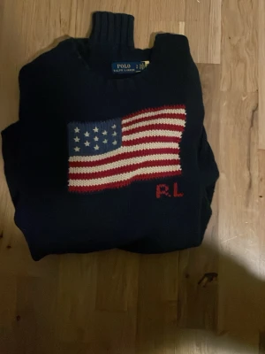 Mörkblå stickad tröja Polo Ralph Lauren - Mörkblå stickad tröja från Polo Ralph Lauren med amerikansk flagga i rött, vitt och blått på bröstet samt RL-broderi. Tröjan har hög krage och klassisk passform. Perfekt för dig som gillar preppy stil och snygga detaljer.