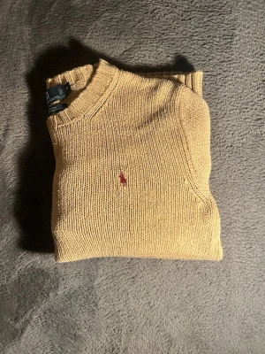 Beige stickad tröja från Polo Ralph Lauren -    ✨ Klassisk stickad tröja från Polo Ralph Lauren ✨  En tidlös och stilren stickad tröja i en mjuk beige nyans. Tröjan har en klassisk passform med rund hals, ribbade muddar och den ikoniska broderade Polo-loggan på bröstet. Perfekt både till vardags och för en mer uppklädd look – lika snygg till jeans som till chinos.