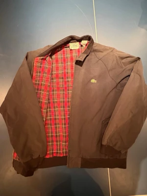 Svart bomberjacka från Lacoste - Klassisk svart bomberjacka från Lacoste med ikonisk krokodillogga på bröstet. Jackan har ribbade muddar och krage samt röd- och grönrutigt innerfoder. Perfekt för dig som gillar stilren design med en twist. Yttermaterialet är bomull.