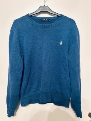 Blå tröja från Polo Ralph Lauren - Klassisk blå stickad tröja från Polo Ralph Lauren med rund halsringning och ribbade muddar. Tröjan har den ikoniska broderade loggan i gult på bröstet och är tillverkad i mjukt material. Perfekt för en stilren och avslappnad look.