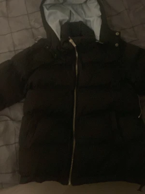 Svart pufferjacka med huva - Säljer en svart pufferjacka  stor huva och ljusblått innerfoder. Jackan har dragkedja framtill och två stora fickor. Perfekt för kalla vinterdagar och riktigt skön att bära. Klassisk design som funkar till det mesta.