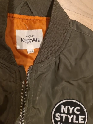 Grön bomberjacka från KappAhl - Säljer en grön bomberjacka från KappAhl med coola NYC-patchar och orange innerfoder. Jackan har dragkedja framtill, ribbade muddar och fickor med knapp. Perfekt för dig som gillar streetstyle och vill sticka ut lite extra.