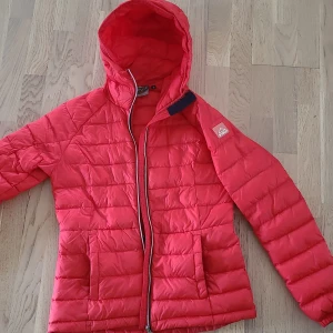 Röd pufferjacka från McKinley, strl 38 - Säljer en röd pufferjacka från McKinley i storlek 38. Jackan har huva, två fickor framtill och dragkedja hela vägen. Quiltad design och sportig look, perfekt för kyliga dagar. Jackan är lätt och smidig att bära.