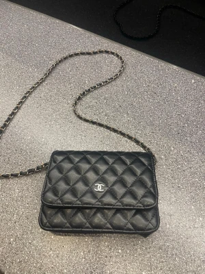 Svart quiltad Chanel handväska - Säljer en svart quiltad handväska från Chanel med klassiskt kedjeband och silverfärgad logga framtill. Väskan är tillverkad i läder och har en tidlös, elegant design med rutmönster. Perfekt accessoar för dig som gillar lyxiga detaljer.