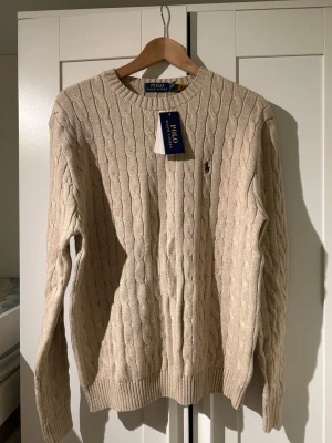 Beige kabelstickad tröja Polo Ralph Lauren - Kabelstickad beige tröja från Polo Ralph Lauren med rund halsringning och ribbade muddar. Tröjan har den klassiska broderade loggan brunt på bröstet och är tillverkad i mjukt material. Perfekt för dig som gillar stilren och tidlös design. Helt ny oanvänd. Priset kan diskuteras. Storlek M