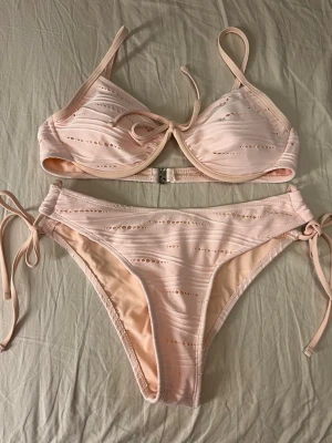 Ljusrosa bikini med knyt och håldetaljer - Supersöt ljusrosa bikini med håldetaljer och knyt i sidorna på underdelen samt framtill på överdelen. Bikinin har tunna axelband och en metallspänne i ryggen. Materialet känns mjukt och stretchigt, perfekt för sommardagar på stranden. Säljer då den är i fel storlek, endast testad aldrig använd