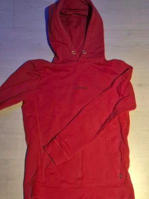 Röd hoodie från Les Deux - Snygg röd hoodie från Les Deux med broderad logga på bröstet. Hoodien har klassisk huva med dragsko och en stor magficka. Tillverkad i mjukt material som känns skönt mot huden. Perfekt för en avslappnad och trendig stil.