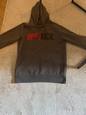 Hoodrich hoodie - Snygg grå hoodie från Hoodrich med stor huva och magficka. Tryck på bröstet i rött och svart där det står 'HOODRICH.'. Perfekt för en streetwear-look och riktigt skön att ha på sig. Materialet känns mjukt och bekvämt.