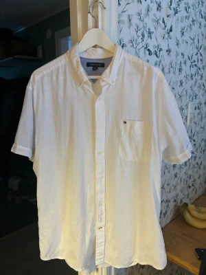 Vit linneskjorta från Tommy Hilfiger - Klassisk vit skjorta från Tommy Hilfiger i svalt linne. Skjortan har korta ärmar, bröstficka med diskret logga och knappar framtill. Perfekt för en clean och stilren look.