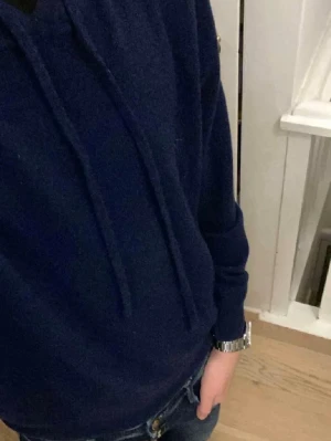 Kashmir Hoodie - Säljer en mörkblå kashmir hoodie med klassisk huva och dragsnören. Hoodien är tillverkad i mjukt material och har ribbade muddar vid ärmslut och nederkant. Perfekt för en avslappnad och stilren look. Tröjan har ett väldigt litet hål men inget som märks.