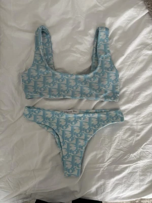 Blåvit bikini från Christian Dior - Säljer en snygg blåvit bikini från Christian Dior med ett ikoniskt mönster. Överdelen har breda axelband och underdelen är högt skuren. Perfekt för sommaren och strandhäng. Materialet känns mjukt och stretchigt.