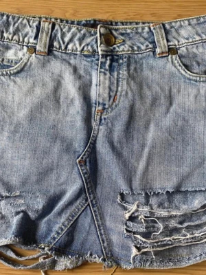 Ljusblå  jeanskjol  - Trendig ljusblå jeanskjol med råa kanter och slitna detaljer framtill. Kjolen har klassisk femficksmodell, knapp- och dragkedjestängning samt fransig nederkant för en avslappnad look. Red Label. 