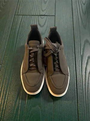 Mörkgröna Luboutines  sneakers med röd sula - Snygga mör sneakers med vit kant och ikonisk röd sula. Skorna har en stilren design med rundad tå och matchande mörkgrå snören. Materialet ser ut att vara textil eller mocka för en modern look. Perfekta för dig som vill sticka ut med detaljer.