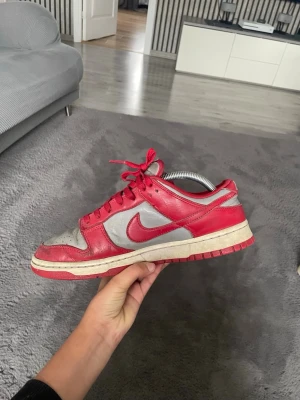 Nike Dunk Low röd och grå sneaker - Säljer ett par Nike Dunk Low sneakers i rött och grått läder med klassisk röd swoosh. Skorna har vita och röda detaljer, lågt skaft och snörning. Perfekta för dig som gillar streetwear och vill ha en ikonisk modell i din samling.