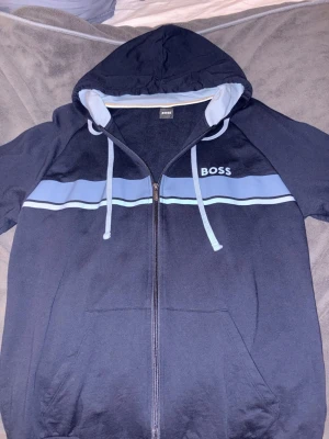 Marinblå hoodie från BOSS - Snygg marinblå hoodie från BOSS med dragkedja framtill, ljusblå och vit rand över bröstet samt justerbar huva med snören. Hoodien har fickor framtill och en sportig look. Perfekt för dig som gillar stilrena och bekväma plagg.