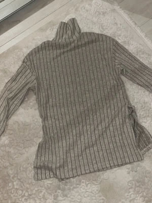 Grå ribbstickad polotröja - Mysig grå ribbstickad polotröja med lång ärm och hög krage. Tröjan har en avslappnad passform och är tillverkad i ett mjukt material som känns skönt mot huden. Perfekt för lager på lager och en stilren look.