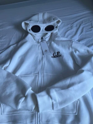 C.P. Company hoodie - Säljer en vit hoodie från C.P. Company med dragkedja och ikoniska goggles i huvan. Hoodien har broderad logga på bröstet och är tillverkad i mjukt material. Perfekt för dig som vill sticka ut med en unik detalj i streetwear-stil.