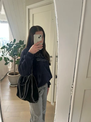 Svart Stella McCartney liknande väska - Snygg svart handväska med kedjedetaljer i silver runt kanterna och som axelrem. Väskan har en mjuk, slouchy form. Perfekt för dig som vill ha en trendig och rymlig väska med coola detaljer.