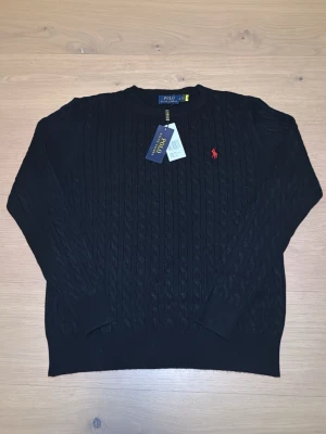 Svart kabelstickad tröja Polo Ralph Lauren - Kabelstickad svart tröja från Polo Ralph Lauren med rund halsringning och röd broderad logga på bröstet. Tröjan har långa ärmar och ribbade muddar vid ärmslut och nederkant. Perfekt för dig som gillar klassisk och stilren design. Storleken är L men passar storlek S