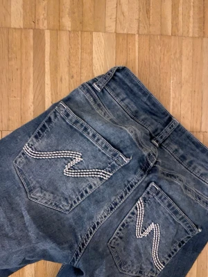 Blå Nudie Jeans med broderade fickor - Säljer ett par klassiska blå jeans från Nelly med deras ikoniska broderade vågmönster på bakfickorna. Jeansen är low waist bootcut i storlek 34. Pris kan såklart diskuteras 