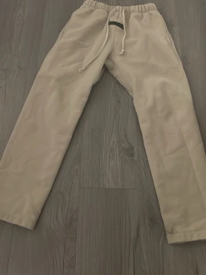 Beige mjukisbyxor från Essentials - Beiga mjukisbyxor från Essentials Fear of God med broderad logga på benet. Byxorna har raka ben, resår och dragsko i midjan för skön passform. Perfekta för en chill och avslappnad stil.