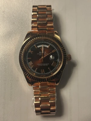 Guldfärgad klocka Day-Date - Snygg guldfärgad Day-Date med mörkbrun urtavla och dag- samt datumvisning. Klockan har en klassisk länkarmband i metall och markerade index. Perfekt accessoar för dig som vill sticka ut med stil.