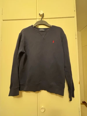 Mörkblå sweatshirt från Polo Ralph Lauren - Mörkblå sweatshirt från Polo Ralph Lauren med klassisk rund halsringning och röd broderad logga på bröstet. Tröjan har ribbade muddar vid ärmslut och nederkant. Perfekt för en avslappnad och stilren look.