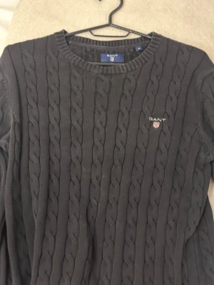 Mörkgrå kabelstickad tröja GANT - Snygg mörkgrå kabelstickad tröja från GANT med rund halsringning och broderad logga på bröstet. Tröjan har klassiskt stickat mönster och ribbade muddar vid hals, ärmslut och nederkant. Perfekt för lager-på-lager och stilren look.