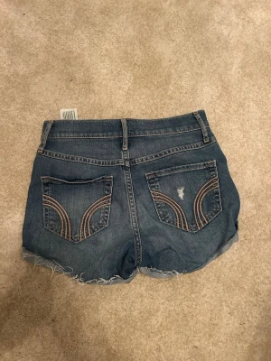 Blå jeansshorts från Hollister - Snygga blå jeansshorts från Hollister med slitna detaljer och råa kanter. Shortsen har fem fickor, bälteshällor och dekorativa sömmar på bakfickorna. Perfekta för varma dagar och en avslappnad stil.