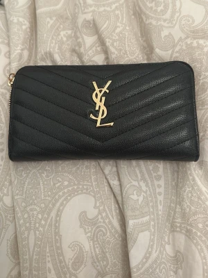 Svart YSL plånbok i läder - Snygg svart plånbok från Saint Laurent med klassiskt YSL-logo i guld framtill. Plånboken är tillverkad i strukturerat läder med quiltad design och dragkedja längs kanten. Perfekt för dig som gillar lyxiga accessoarer och stilren look.