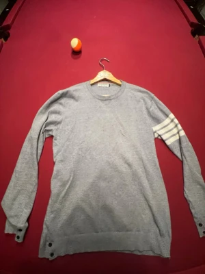 Grå stickad tröja Thom Browne - Snygg grå stickad tröja från Thom Browne med rund halsringning och fyra vita ränder på ena ärmen. Tröjan har långa ärmar och knappar vid ärmslut och nederkant. Perfekt för dig som gillar stilrena detaljer och exklusiva märken.