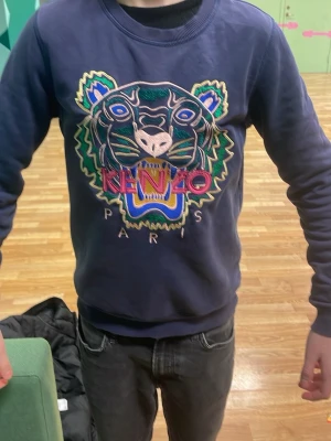 Kenzo tröja kostar 2700kr Ny!!! - Säljer en blå hoodie från Kenzo med ikoniskt broderat tigerhuvud och färgglada detaljer på bröstet. Tröjan har rund halsringning och ribbade muddar. Perfekt för dig som vill sticka ut med en streetstyle-look. Kostar 2700kr Ny 