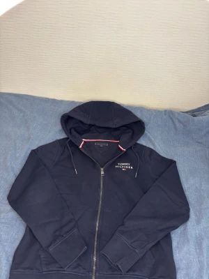 Mörkblå hoodie från Tommy Hilfiger - Snygg mörk blå hoodie från Tommy Hilfiger med dragkedja framtill och justerbar huva. Diskret logga broderad på bröstet och klassiska Hilfiger-detaljer i rött och vitt vid kragen. Är i bra skick.