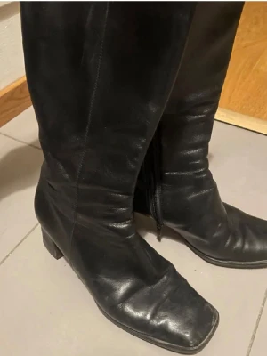 Svarta läderstövlar med klack - Klassiska svarta stövlar i äkta läder från Vero Cuoio. Modellen har fyrkantig tå, medelklack och dragkedja på insidan. Perfekta till höst- och vinterlooken, snygga till både jeans och kjol.