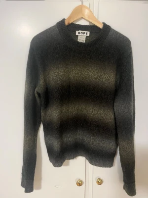 Gråmelerad stickad tröja från HOPE - Stickad tröja från HOPE i gråmelerad ton med breda horisontella ränder i olika nyanser av grått och brunt. Tröjan har rund halsringning, långa ärmar och ribbade muddar vid ärmslut och nederkant. Perfekt för lager på lager och en avslappnad stil.