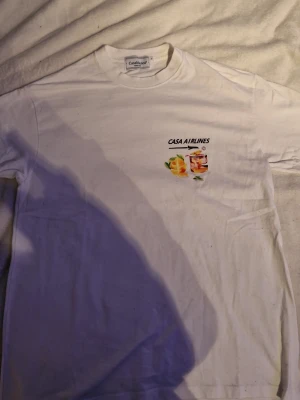 Vit Casa Airlines t-shirt med tryck - Vit t-shirt från Casablanca med coolt färgglatt tryck av drink och apelsiner på bröstet samt stort motiv på ryggen med flygplansfönster, hav och solnedgång. Rund hals och korta ärmar. Perfekt för dig som gillar unika prints och streetwear vibes.