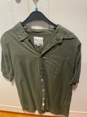 Grön skjorta från 157 med kort ärm - Stilren grön skjorta från 157 med korta ärmar och klassisk krage. Skjortan har en bröstficka och knäppning framtill. Tillverkad i mjuk bomull som känns skön mot huden. Perfekt för en avslappnad och enkel look.