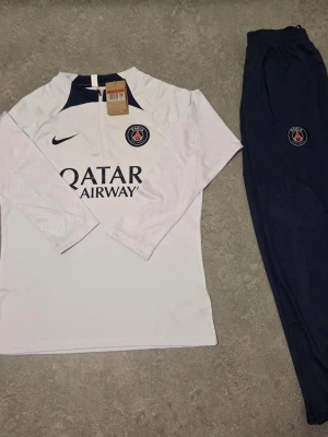 PSG träningsset från Nike - Snyggt PSG träningsset med långärmad vit tröja och mörkblå byxor. Tröjan har dragkedja vid halsen, PSG-logga och Nike Swoosh på bröstet samt Qatar Airways-tryck. Byxorna är mörkblå med PSG-emblem. Perfekt för fotbollsträning eller chill.