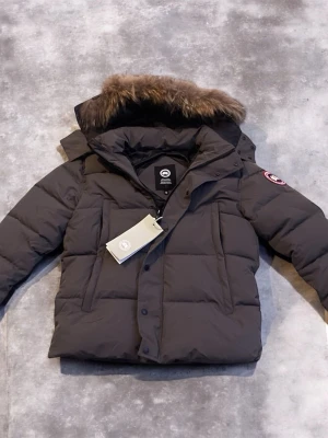   Canada Goose jacka - Snygg mörkgrå dunjacka från Canada Goose med stor huva och fuskpälsdetalj. Jackan har klassisk patch på ärmen, dragkedja och knappar framtill samt flera praktiska fickor. Perfekt för kalla vinterdagar och riktigt varm.
