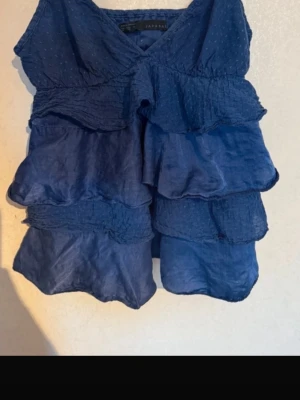 Blå volangtop från Zara Basic - Snygg blå topp från Zara Basic med flera lager volanger och v-ringad hals. Toppen är ärmlös och har en luftig, flowy passform som ger en cool och avslappnad vibe. Perfekt för dig som gillar detaljer och vill sticka ut lite extra.